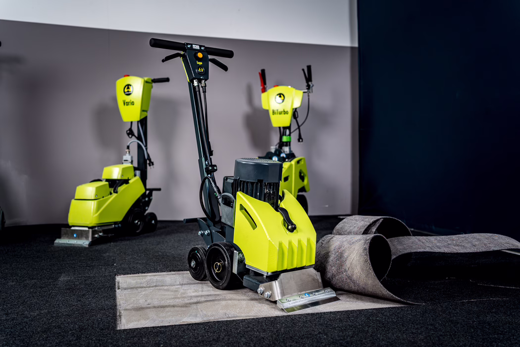WOLFF_Application Image_Lupo_2024-04 (20) A bright yellow-green industrial carpet care machine stands in the foreground of an indoor workshop or showroom. The unit features a black handlebar, visible electric motor housing, two small black wheels and a stainless-steel suction/cleaning head at the front. A section of dark grey commercial carpet has been peeled back and rolled to the right, exposing the lighter backing beneath. Two additional machines in the same high-visibility color are positioned in the background against a two-tone grey wall; one displays the label 'Vario' and the other 'BiTurbo'. The scene is well-lit with a focused, slightly dramatic studio look, the floor covered in low-pile dark carpet. No people are visible; the composition emphasizes the equipment, carpet removal and maintenance context.