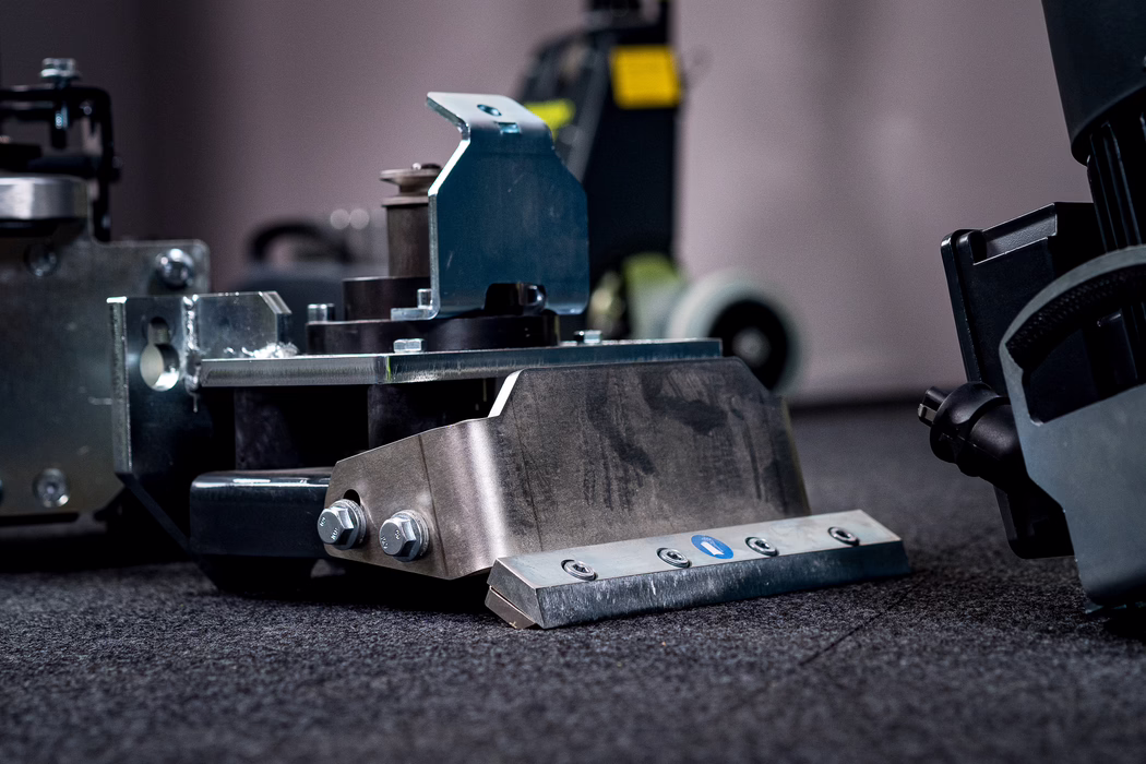 WOLFF_Application Image_Lupo_2024-04 (5) Close-up of a heavy metal industrial component placed on a dark, textured carpet. In the foreground a sturdy stainless steel bracket with two large hex bolts is attached to a beveled metal scraping blade fixed by four countersunk screws; a small blue circular sticker is visible on the blade. The metal surfaces show fine machining marks, slight wear and faint oil or grease smudges. Behind the main part, other machine elements and a caster wheel are visible but blurred by a shallow depth of field. Cool, directional lighting emphasizes the metallic textures, edges and reflections, and the low-angle composition highlights the component's solid, utilitarian construction.